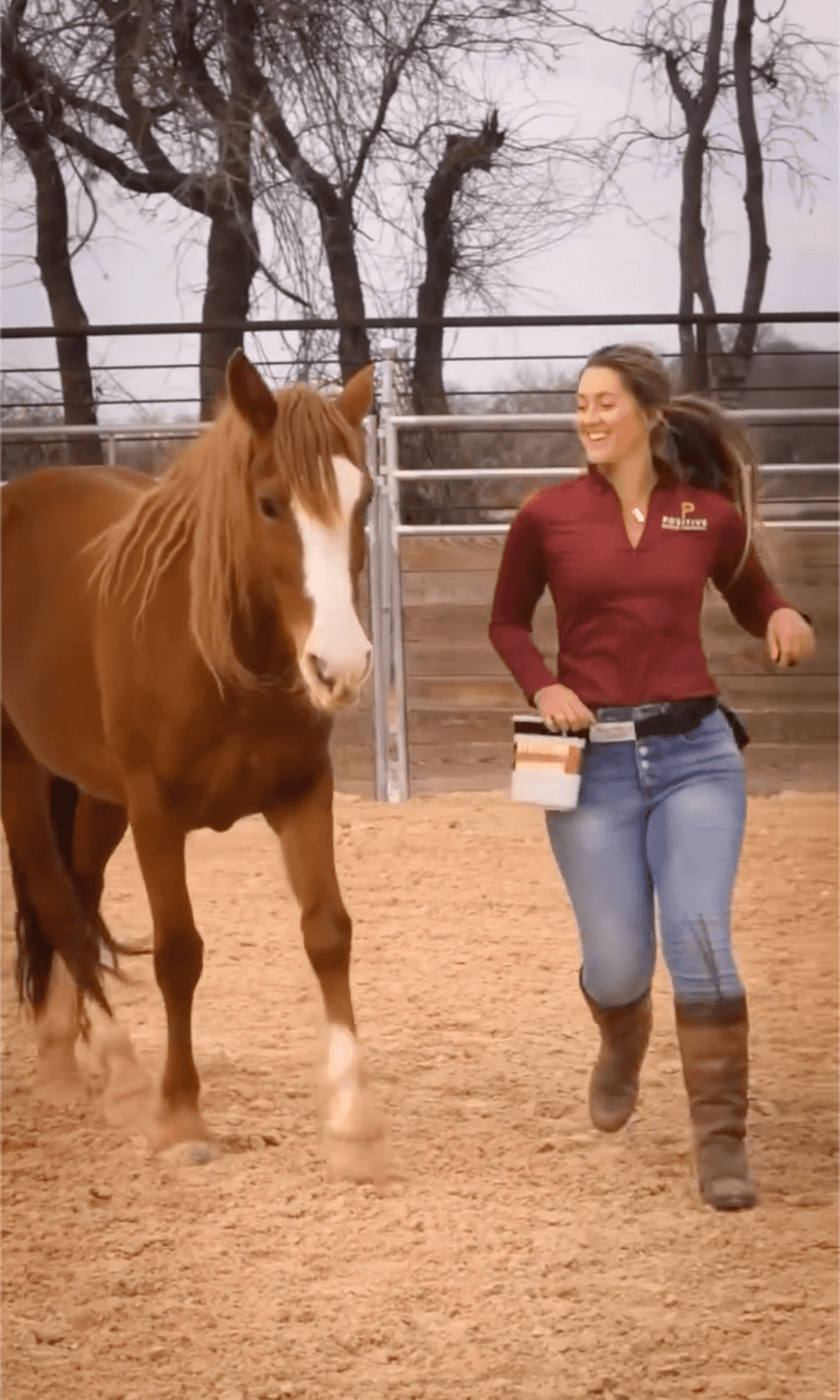 Marina smiling as she walks beside a chestnut horse at liberty in a sandy arena
